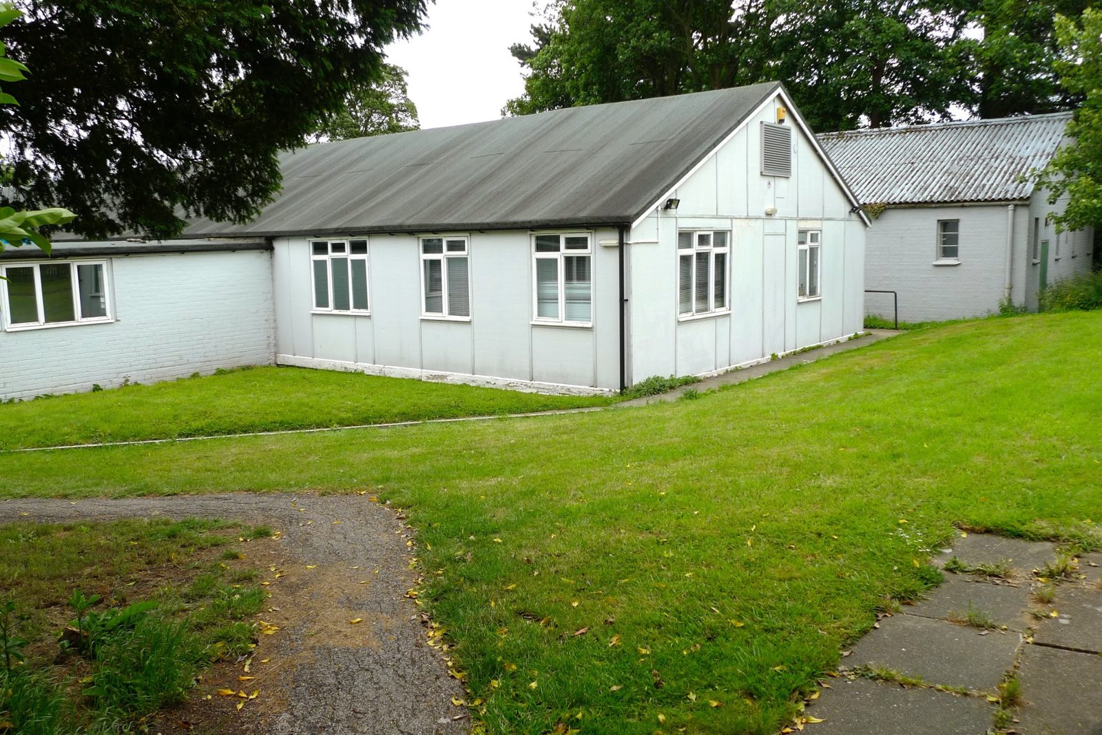 A single-storey white prefabricated hut with a grey pitched roof, on a lawn among trees.